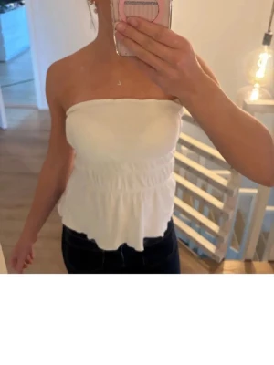 Vit bandeau tubtopp - Säljer en vit bandeau topp från bikbok i storlek xs med smockad midja och volangkant nedtill. Toppen är axelbandslös och har en mjuk, stretchig passform som sitter snyggt på kroppen. Perfekt till jeans eller kjol för en fräsch look💞
