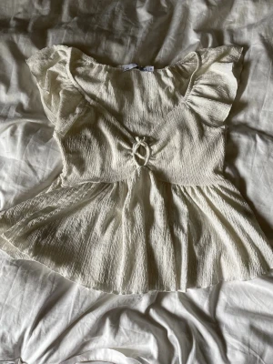 Vit topp med volangärmar - Supersöt vit topp med volangärmar och en snygg knytdetalj framtill. Toppen har en lätt rynkad struktur och är tillverkad i ett mjukt, luftigt material som passar perfekt till sommaren. Den har en feminin siluett och öppen ringdetalj vid bysten.