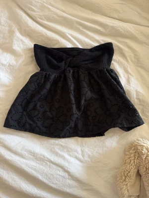 Svart axelbandslös broderad topp - Säljer en svart axelbandslös topp med broderade blommor och smockad överdel. Toppen har en luftig passform och är perfekt för varma dagar. Den är både söt och trendig med sin bandeau-stil och fina detaljer.