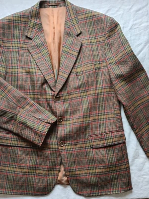 Rutig kavaj i ullmix - Snygg rutig kavaj med klassiskt mönster i grönt, brunt och rött. Tvåknäppt modell med breda slag, tre fickor fram och helfodrad insida i beige. Perfekt för dig som gillar vintagevibbar och tidlös stil. Materialet känns som en ullmix.