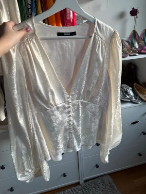 Vit glansig blus från Bik Bok - Stilren vit blus från Bik Bok i glansigt, mönstrat tyg med djup V-ringning och dekorativa knappar framtill. Blusen har långa, puffiga ärmar och lätt figursydd midja för en feminin siluett. Perfekt för att lyfta vilken outfit som helst.