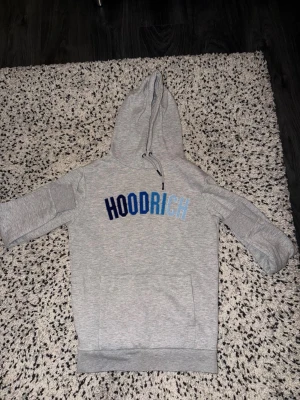 Grå HOODRICH hoodie med blå text - Snygg grå hoodie från HOODRICH med stor broderad logga i olika blå nyanser på bröstet. Klassisk känguruficka framtill och justerbar huva med dragsko. Perfekt för en chill och street-inspirerad look. Mjuk bomullsblandning för skön känsla.