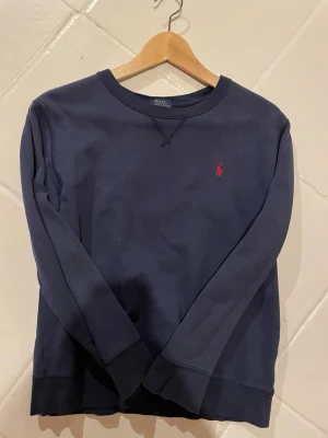 Mörkblå sweatshirt från Polo Ralph Lauren - Klassisk mörkblå sweatshirt från Polo Ralph Lauren med rund halsringning och den ikoniska röda logotypen broderad på bröstet. Tröjan har långa ärmar och ribbade muddar vid ärmslut och nederkant. Perfekt för en avslappnad och stilren look.