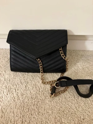 Svart axelväska med kedja - Snygg svart axelväska i quiltat konstläder med guldfärgad kedjerem och kuvertformad lock. Väskan har ett stilrent mönster i v-format och passar perfekt till en trendig look. Har små fläckar på insidan som jag tyvärr inte kunnat få bort