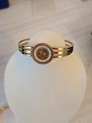 Guldfärgat armband med myntdetalj - Snyggt guldfärgat armband med tre smala band och en rund myntdetalj i mitten. Myntet har ett motiv och text runtom, och armbandet har en elegant, stilren design. Perfekt accessoar för att lyfta din outfit med en touch av lyx.kopar