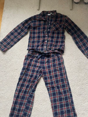 Rutig pyjamas från Gina Tricot - Mysig tvådelad pyjamas från Gina Tricot i blått, rött och grönt rutmönster. Skjortan har långa ärmar, knappar framtill och bröstficka. Byxorna har resår i midjan och en svart satinrosett. Perfekt för chillkvällar hemma.