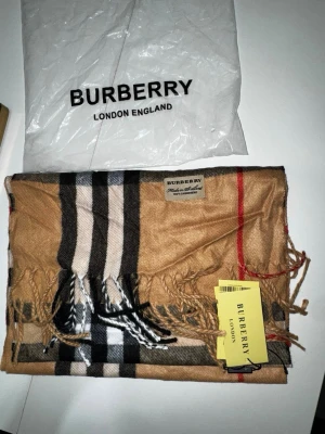 Beige Kashmir halsduk från Burberry  - Klassisk beige halsduk från Burberry i 100% kashmir med ikoniskt rutmönster i svart, vitt och rött. Halsduken har fransar i kanterna och kommer med originaltaggar och påse. Perfekt accessoar för att lyfta din outfit med en tidlös touch. 