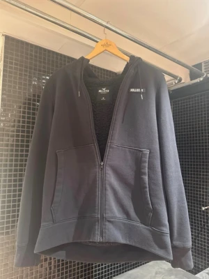 Svart hoodie med dragkedja från Hollister - Svart hoodie från Hollister med dragkedja framtill, två stora fickor och huva. Insidan är fodrad med mjukt material för extra komfort. Klassisk och enkel design med liten logga på bröstet. Perfekt för en avslappnad stil.