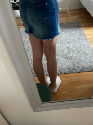 Blå jeanskjol med rå kant - Säljer en blå jeanskjol med rå, fransig kant och klassiska fickor bak. Kjolen har en tight passform och är kort, perfekt för dig som gillar en avslappnad och cool stil. Materialet är denim och färgen är mörkblå med ljusare slitningar.