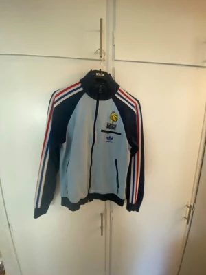 Adidas FFF track jacket blå/vit - Snygg Adidas track jacket i blått och vitt med röda detaljer. Jackan har FFF-logga och Adidas-märke på bröstet samt texten 'FRANCE' på ryggen. Dragkedja framtill, ståkrage och randiga ärmar i rött, vitt och blått. Perfekt för sport eller streetwear.