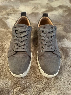 Cristian Louboutin  - Stilrena sneakers i grå mocka med matchande grå skosnören och vit gummisula. Låg modell med diskret design och ljusbrunt innerfoder. Perfekta för dig som gillar en clean och enkel look.