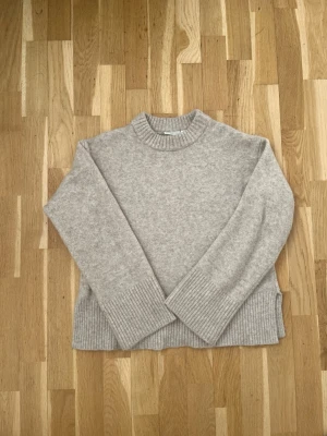 Ljusgrå stickad tröja med ribbade detaljer - En ljusgrå stickad tröja med ribbade muddar vid ärmslut, nederkant och halsringning. Tröjan har en rak och något boxig passform med lång ärm och rund hals. Perfekt för lager-på-lager och enkel att matcha med jeans eller kjol.