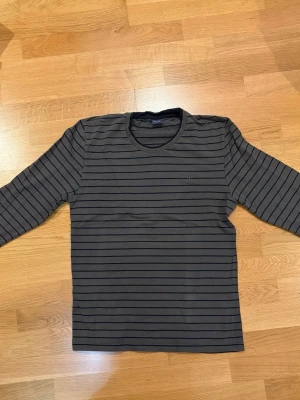 Randig långärmad tröja från Gant - Snygg långärmad tröja från Gant i mörkgrått med tunna svarta horisontella ränder. Tröjan har rund halsringning och en enkel, avslappnad passform. Perfekt för en stilren och casual look.