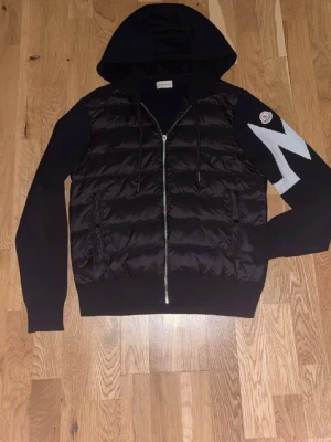 Moncler cardigan  - Snygg svart moncler cardigan i storlek M med quiltad front och stickade ärmar. Jackan har dragkedja, huva med snörning och ett stor vitt M på ärmen samt Moncler logga. Perfekt för dig som vill ha en skön och trendig look. Svarar alla meddelanden direkt och fraktar alltid samma dag som köp:)