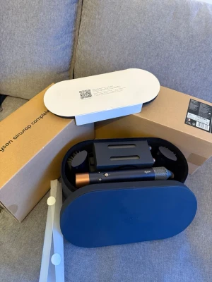 Dyson Airwrap Complete Long hårstyler - Dyson Airwrap Complete Long hårstyler i elegant svart och kopparfärg. Levereras i en oval, hård förvaringsbox med flera stylingtillbehör. Perfekt för att skapa olika frisyrer med hjälp av luft och värme. Materialet är plast och metall, och designen är modern och stilren.