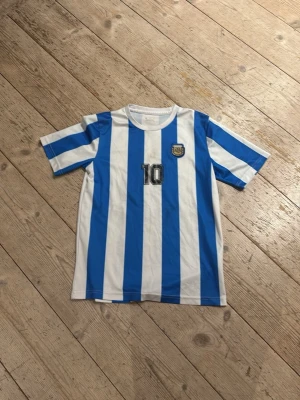 Argentina Maradona tröja nr 10 - Säljer en klassisk fotbollströja med blå och vita vertikala ränder, inspirerad av Argentinas landslag. Nummer 10 och namnet Maradona på ryggen. Tröjan har korta ärmar och är tillverkad i ett lätt, syntetiskt material. Perfekt för fotbollsträning eller som supporterplagg.