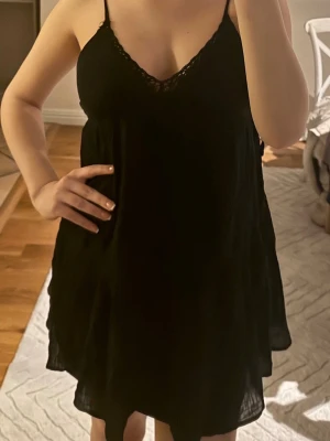 Svart klänning med spets - Svart klänning med tunna justerbara axelband och v-ringad hals med spetsdetalj. Lätt och flödande material som känns bekvämt mot huden. Perfekt för dig som vill ha något enkelt och stilrent. Använts en gång tidigare.