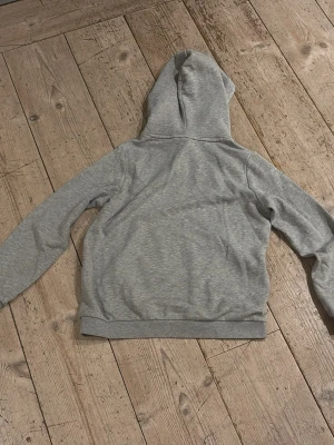 Grå hoodie från Sail Racing - Snygg grå hoodie från Sail Racing med svart logga på bröstet. Klassisk modell med huva och känguruficka framtill. Tillverkad i mjukt bomullsmaterial som känns skönt mot huden. Perfekt för en avslappnad och sportig stil.