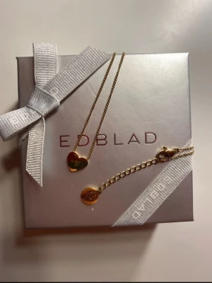 Guldfärgat hjärthalsband från Edblad - Snyggt halsband från Edblad i guldfärgad metall med ett hjärtformat hänge. Kedjan är tunn och elegant, perfekt för att ge en stilren touch till din outfit. Levereras i original ask med vit rosett.