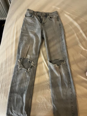 Ljusblå ripped jeans från gina tricot - Säljer ett par ljusblå jeans från gina tricot jeans med raka ben och stora slitningar över båda knäna. Klassisk femficksmodell i denim med coolt, slitet utseende. Perfekta för en avslappnad streetstil.