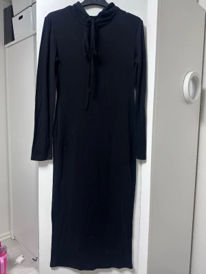  Svart klänning med luva - En svart långklänning med långa ärmar och luva. Klänningen har en enkel, rak passform och är tillverkad i ett mjukt material som känns bekvämt mot huden. Perfekt för dig som gillar stilrena och minimalistiska plagg.