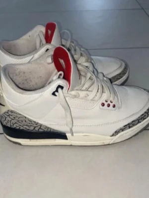 Jordan 3 White Cement - Säljer ett par Nike Air Jordan 3 White Cement sneakers med vit ovandel i läder, ikoniska elefantmönstrade detaljer vid tå och häl, samt röda och svarta inslag. Skorna har klassisk hög komfort och retrostil med synlig Air-dämpning i sulan.