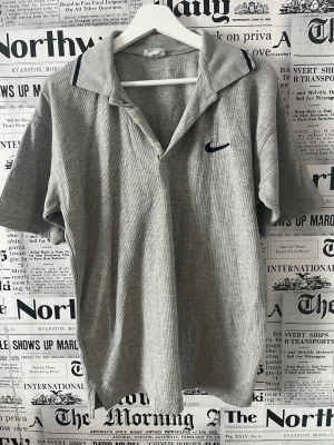 Grå ribbad pikéskjorta från Nike - Grå ribbad pikéskjorta från Nike med korta ärmar och klassisk krage. Skjortan har knappar framtill och en mörkblå Nike-logga på bröstet. Perfekt för en avslappnad och sportig stil. Den har ett litet hål se bild och måttet kan ni se på bilderna med 