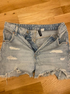 Ljusblå denimshorts från Bershka - Säljer ett par ljusblå denimshorts från Bershka med råa, fransiga kanter och slitna detaljer framtill. Klassisk femficksmodell med bälteshällor och knappgylf. 