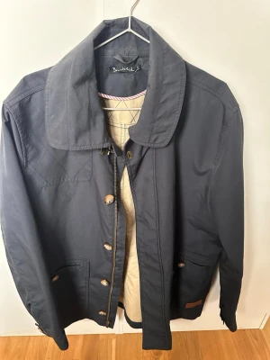 Mörkblå jacka från Bondelid - Klassisk mörkblå jacka från Bondelid med stora fickor, bred krage och både dragkedja och knappar framtill. Jackan har quiltade detaljer på axlarna och är perfekt för dig som gillar tidlös stil.