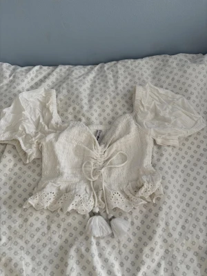 Vit blus med volang och tofsar  - Supersöt kort vit blus med puffärm, snörning framtill och volangkant med broderade detaljer. Blusen har en v-ringad urringning och dekorativa tofsar. Perfekt för en somrig och söt look💗☀️.