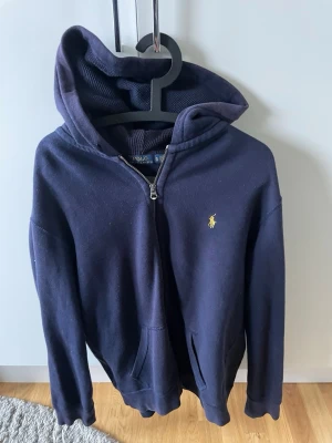Mörkblå hoodie från Polo Ralph Lauren - Mörkblå hoodie med dragkedja från Polo Ralph Lauren. Klassisk gul logga broderad på bröstet, två fickor framtill och skön huva. Tillverkad i mjukt bomullsmaterial som känns riktigt bekvämt. Perfekt för en avslappnad och stilren look.