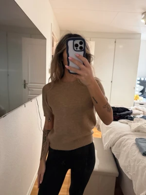 Brun stickad tröja med kort ärm - Säljer en enkel och stilren brun stickad tröja med korta ärmar och rund hals. Tröjan har ribbade muddar vid ärmslut och nederkant, vilket ger en snygg passform. Perfekt att matcha med jeans eller kjol för en avslappnad look.