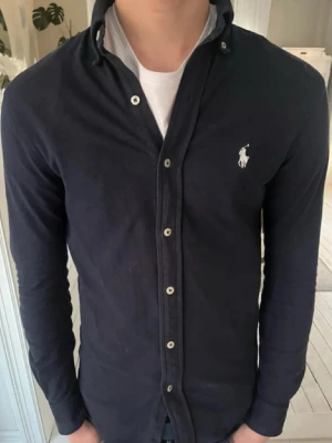 Marinblå skjorta från Ralph Lauren - Klassisk marinblå skjorta från Ralph Lauren med vit broderad logga på bröstet. Skjortan har lång ärm, knappar framtill och är gjord i mjuk bomull. Perfekt för en clean och stilren look. Säljs då den är förliten!