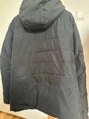 Svart pufferjacka från Peak Performance - Säljer en svart pufferjacka från Peak Performance med huva och flera praktiska dragkedjefickor framtill. Jackan har quiltad design och är perfekt för kalla vinterdagar. Materialet är slitstarkt och håller värmen bra. Snygg och funktionell jacka för dig som vill hålla stilen i vinter.