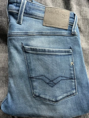 Ljusblå Anbass jeans från Replay - Snygga ljusblå slim fit jeans från Replay med klassisk femficksmodell och diskret slitning framtill. Jeansen har en läderpatch med Replay-logga bak i midjan och mörkblå sömmar på bakfickan. Perfekta för en avslappnad och trendig look. Längd 34 och midja 34. Skick 9,5/10
