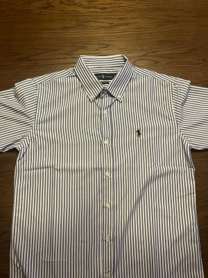 Blårandig skjorta från Ralph Lauren - Klassisk blårandig skjorta från Ralph Lauren med button-down krage och broderad logga på bröstet. Skjortan har vita knappar och är tillverkad i bomull. Perfekt för en stilren och tidlös look. Skjortan är i storlek M 