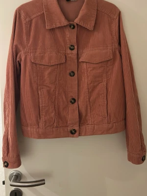 Rosa jeansjacka med fickor - Snygg rosa jeansjacka med klassisk krage och stora bruna knappar. Två bröstfickor med lock och raka sömmar framtill. Jackan har långa ärmar och en rak passform som ger en avslappnad look. Jackan är nu ett par år, men är lite sliten i modellen, jackan är väl omhändertagen och har inga defekter. 