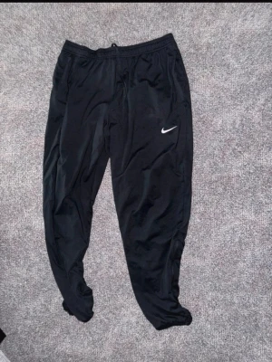 Svarta Nike Dri-FIT träningsbyxor - Säljer ett par svarta träningsbyxor från Nike med Dri-FIT-material som transporterar bort fukt. Byxorna har resår i midjan, smal passform och en vit Nike-logga på benet. Perfekta för gymmet eller löpning.