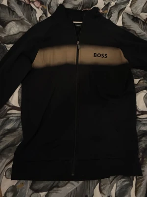 Svart zip hoodie från BOSS - Snygg svart hoodie från BOSS med dragkedja framtill och en beige panel över bröstet där BOSS-loggan är tryckt. Hoodien har långa ärmar och ribbade muddar. Perfekt för en clean och stilren look. Materialet känns mjukt och bekvämt.