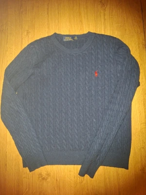 Mörkblå kabelstickad tröja Polo Ralph Lauren - Kabelstickad mörkblå tröja från Polo Ralph Lauren med rund halsringning och röd broderad logga på bröstet. Tröjan har långa ärmar och ribbade muddar vid ärmslut och nederkant. Perfekt för en stilren och klassisk look.