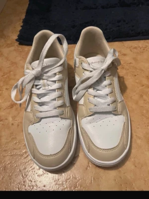 Beige och vita sneakers från Champion - Snygga sneakers från Champion i beige och vitt. Skorna har klassisk låg modell med vita snören och perforerad tå för extra ventilation. Tillverkade i syntetmaterial med stilrena detaljer och bekväm passform. Perfekta för en trendig och avslappnad look.