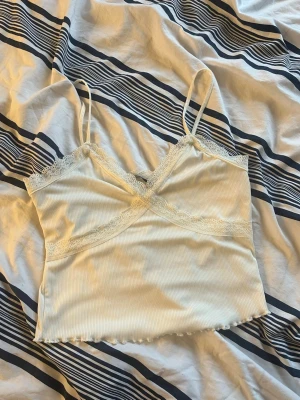 Vit ribbad linnetopp med spets - Söt vit ribbad linnetopp med tunna axelband och spetsdetaljer längs urringningen. Toppen har en croppad passform och vågig kant nertill. Banden kan vara lite långa men säkert lättfixat vid behov. 93% polyester & 7% elastan. Otroligt sällan använd.