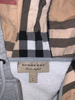 Grå hoodie från Burberry med dragkedja - Snygg grå hoodie från Burberry med dragkedja och klassiskt rutigt foder i huvan. Mjuk insida, två stora fickor framtill och justerbar huva med snören. Perfekt för en chill och stilren look. Den är helt perfekt och inget fel med den ser ut som helt ny