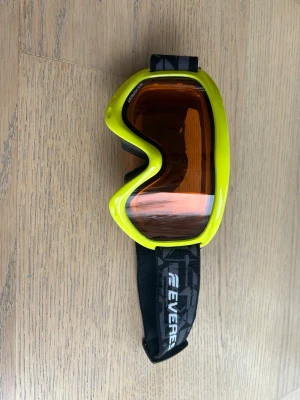 Gula skidglasögon Everest - Coola skidglasögon från Everest med gul ram och orange tonade linser. Justerbart svart band med mönster och logga. Perfekta för skidåkning eller snowboard. Sköna skumkanter för extra komfort och bra passform. 19cm läng från framsidan. 10cm från insidan.