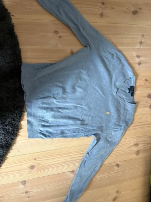 Grå tröja från Polo Ralph Lauren - Snygg grå tröja från Polo Ralph Lauren med klassisk gul broderad logga på bröstet. Tröjan har rund hals och långa ärmar, perfekt för en stilren look. Materialet är mjukt och tunt stickat, vilket gör den bekväm att bära.