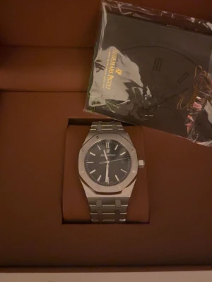 Audemars Piguet Royal Oak klocka silver - Säljer en Audemars Piguet Royal Oak klocka i rostfritt stål med silverfärgad länk och svart urtavla. Klockan har den ikoniska åttakantiga boetten och tydliga indexmarkeringar. Perfekt för dig som gillar exklusiva accessoarer och stilren design.