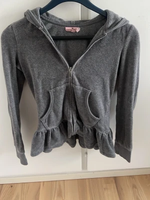 Grå hoodie med volang från Juicy Couture - Mysig grå vintage kofta från Juicy Couture med dragkedja, volangkant nedtill och fickor framtill. Stor logga med glitterdetaljer på ryggen och huva. Tillverkad i mjukt velourmaterial som känns lyxigt och bekvämt.