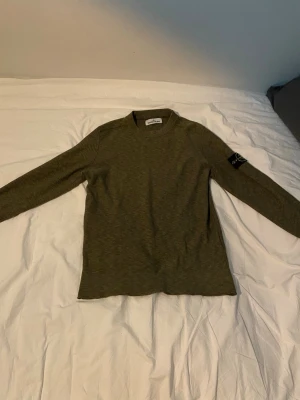 Olivgrön Stone Island tröja - Snygg olivgrön långärmad tröja från Stone Island med den klassiska loggan på vänster ärm. Tröjan har en enkel, rak passform och är tillverkad i mjukt material som passar perfekt till streetwear-stilen.