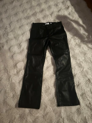 Svarta byxor i fuskskinn - Snygga svarta byxor i fuskskinn med raka ben och klassisk femficksmodell. Byxorna har normal midja och stängs med knapp och dragkedja. Perfekta för dig som vill ha en edgy look.