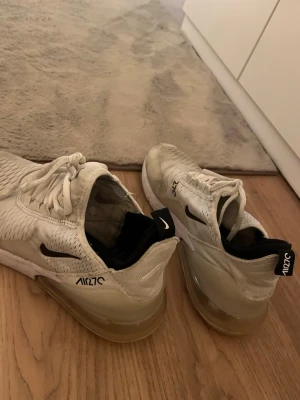 Nike Air Max 270 vita sneakers - Nike Air Max 270 sneakers i vitt med svarta detaljer och stor synlig luftbubbla i hälen. Ovandelen är i mesh för bra ventilation och har klassisk snörning. Snygg och sportig design med AIR270-logga på sidan och bak.